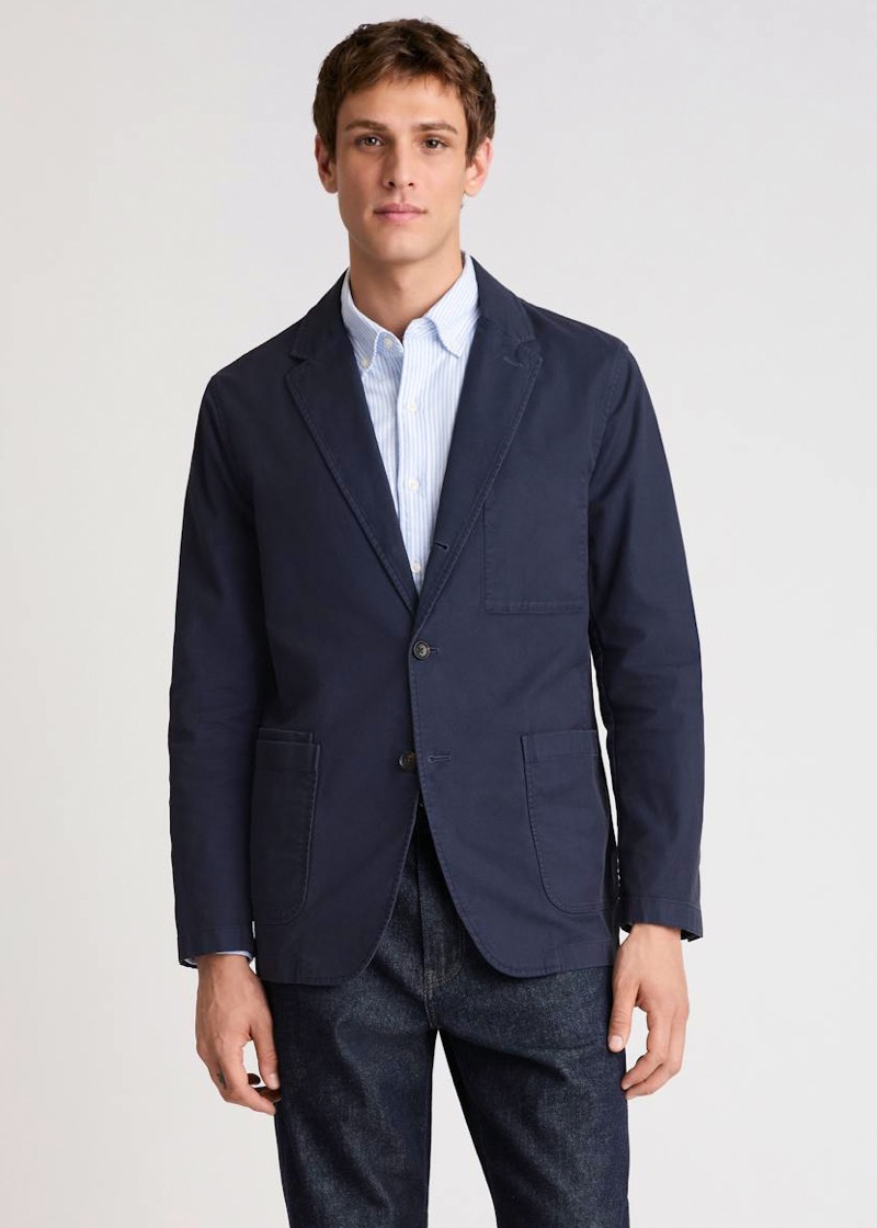 A young man stands in a navy unstructured cotton two-button blazer with patch pockets and soft shoulders over a light blue striped oxford shirt and dark raw denim jeans.
