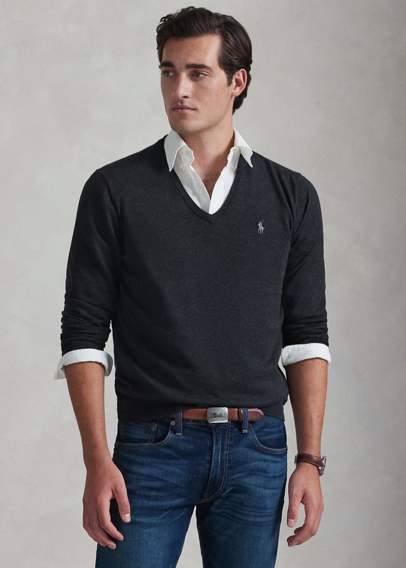 A man with swept-back dark hair wears a charcoal merino V-neck sweater with a small embroidered pony at the chest, layered over a white oxford button-down with collar and cuffs showing, tucked into mid-blue jeans cinched with a tan leather belt.