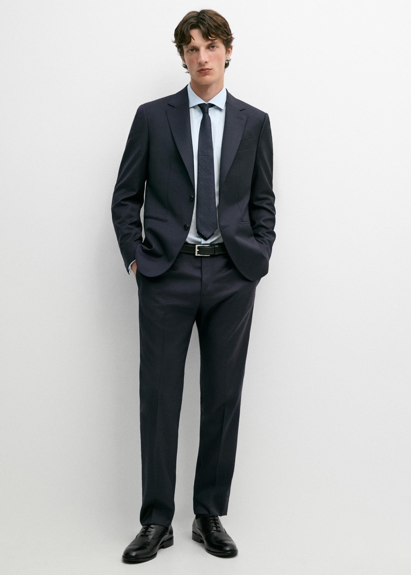 A man wearing a slim-fit dark navy two-button suit with notch lapels and a black leather belt, paired with a light blue dress shirt, a navy patterned silk tie, and black leather Derby shoes.