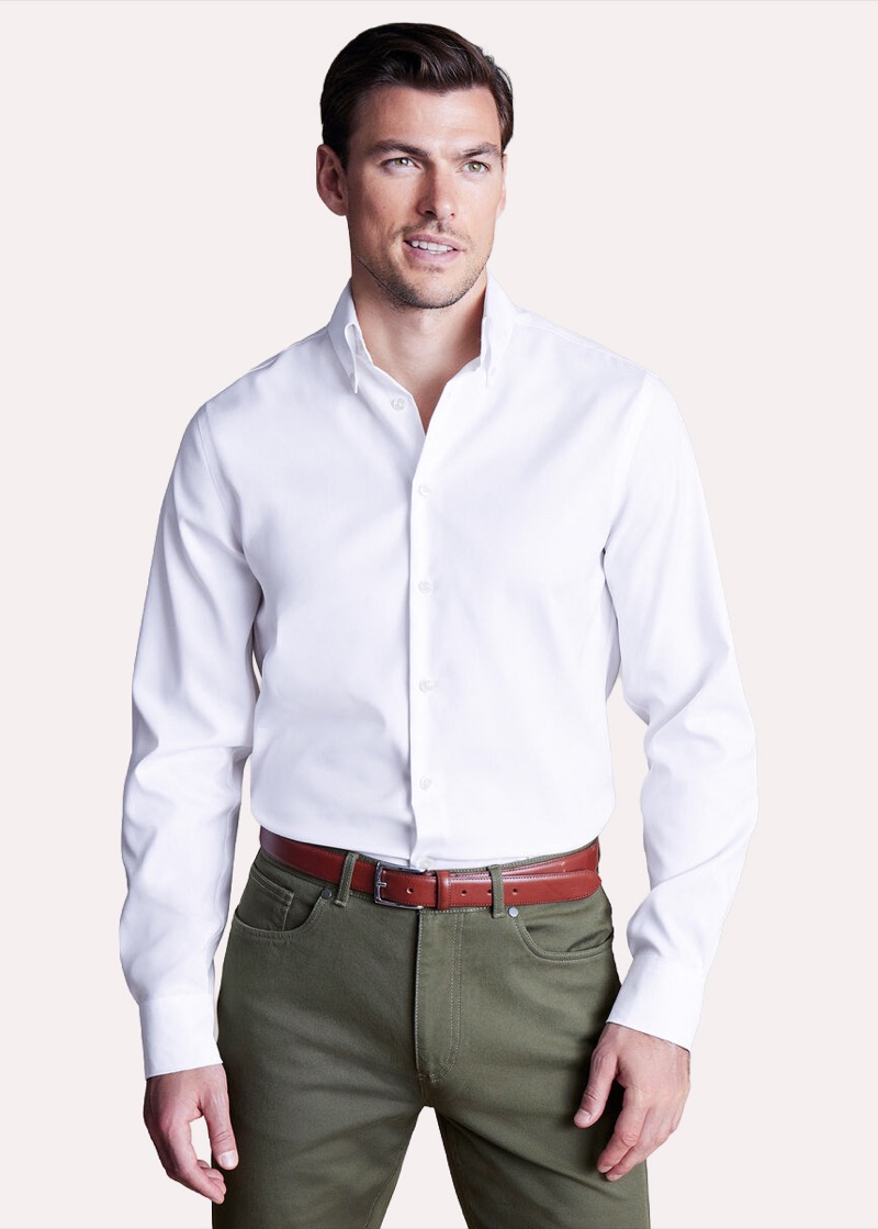 A man with short brown hair stands against a soft pink backdrop in a crisp white non-iron oxford button-down shirt with a soft button-down collar and white buttons, tucked into olive-green five-pocket pants belted with a rich cognac leather strap.