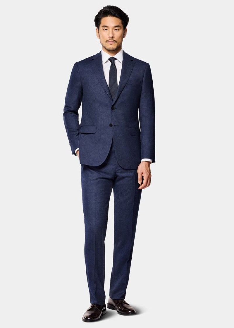 A man wearing a dark navy textured wool two-button suit with notch lapels and flat-front trousers, paired with a white dress shirt, a navy silk tie, and dark brown polished leather Derbies.
