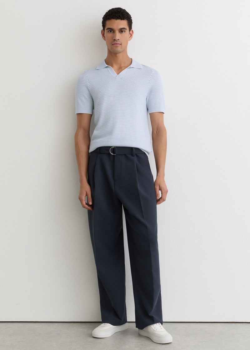 A man in a pale ice-blue knit polo stands in wide-leg navy pleated wool trousers with a D-ring belt at the waist, cuffed at the hem above white minimalist sneakers.