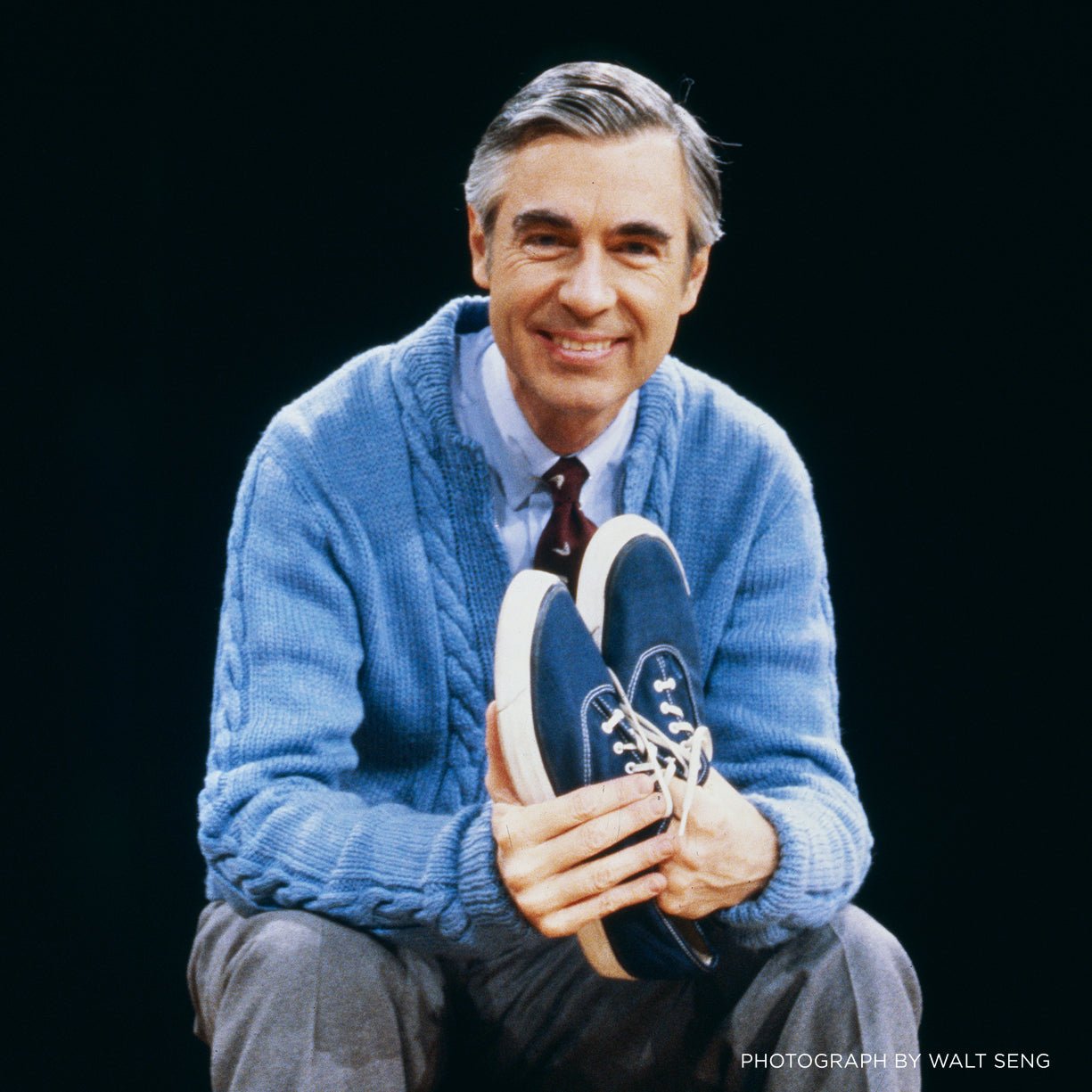 Fred Rogers wears a blue cardigan, shirt and tie while holding a pair of blue canvas sneakers.