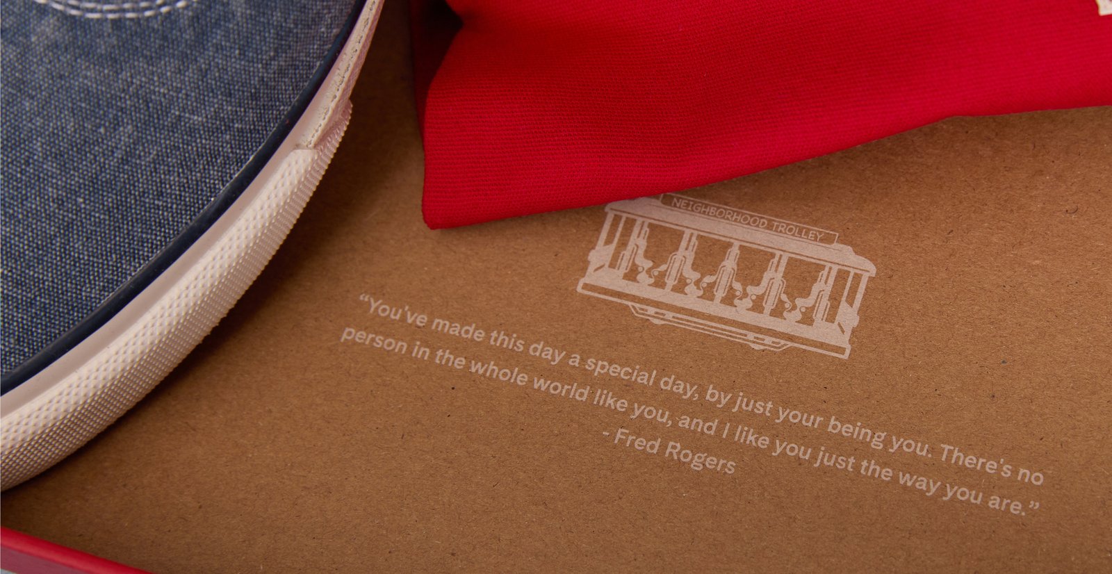 A close-up of a blue canvas sneaker, red knitwear, and collection packaging printed with a trolley illustration and a Fred Rogers message.