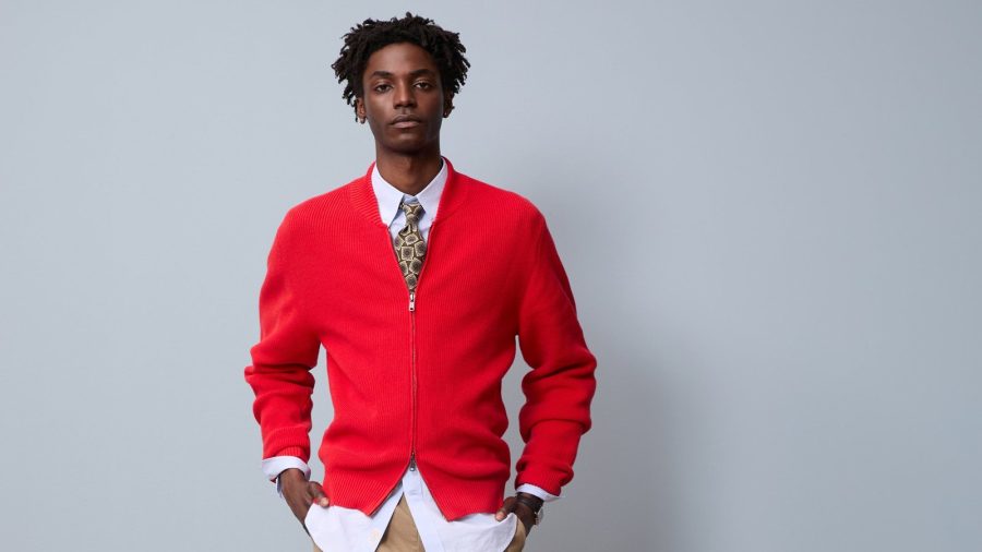 A model wears a red zip-up cardigan, white shirt, patterned tie, cuffed tan trousers, and ecru canvas sneakers beside a wooden armchair.