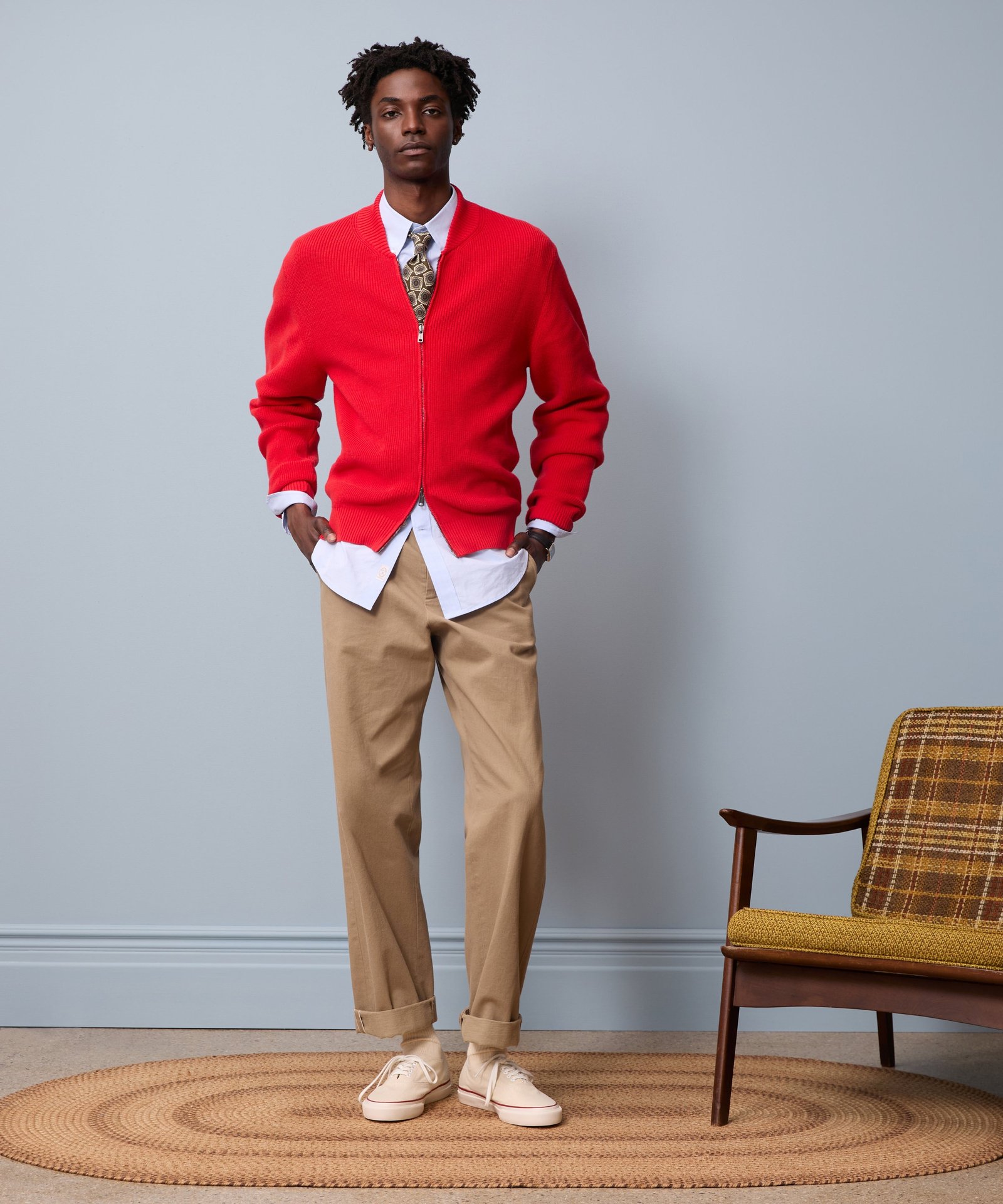 A model wears a red zip-up cardigan, white shirt, patterned tie, cuffed tan trousers, and ecru canvas sneakers beside a wooden armchair.