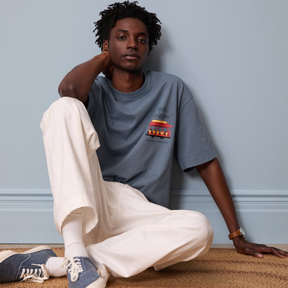 A seated model wears a blue Trolley Tee, cream trousers, white socks, and blue canvas sneakers.