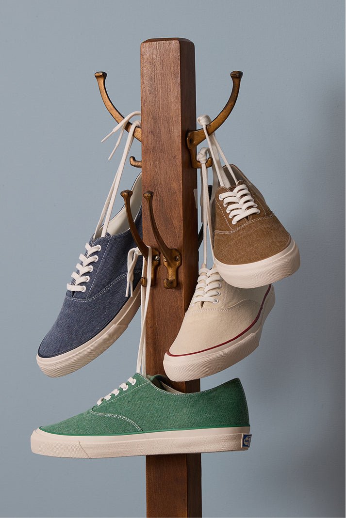 Indigo, brown, ecru, and green canvas CVO sneakers hang by their laces from a wooden coat stand.